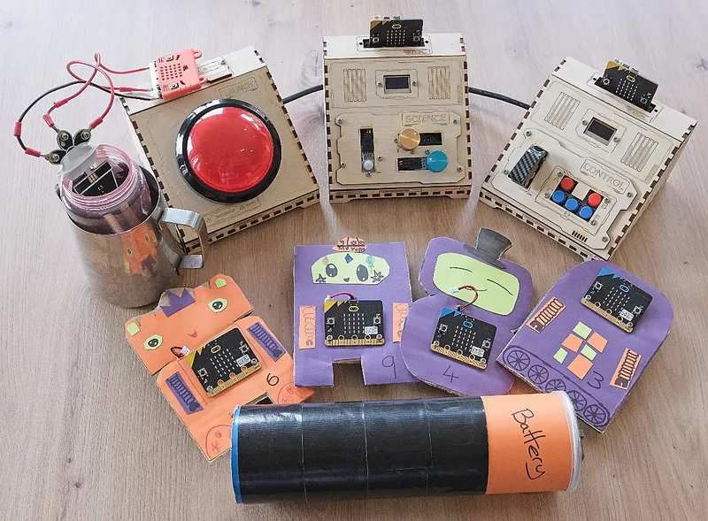 The escape room kit laid out on a table: three laser cut wooden consoles, a big red button, four cardboard robots with micro:bits, a jug and a Pringles tube battery.